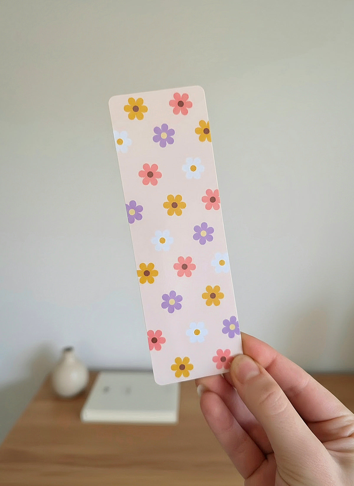 A hand holding a bookmark with simple flower pattern design with yellow, pink, white, and purple daisies on a light pink / cream background.