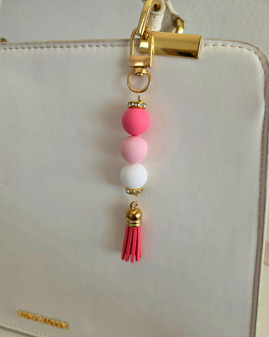 A close-up of a decorative gold-tone zipper pull charm with pink, light pink, and white beads and a pink tassel hanging on a white handbag.