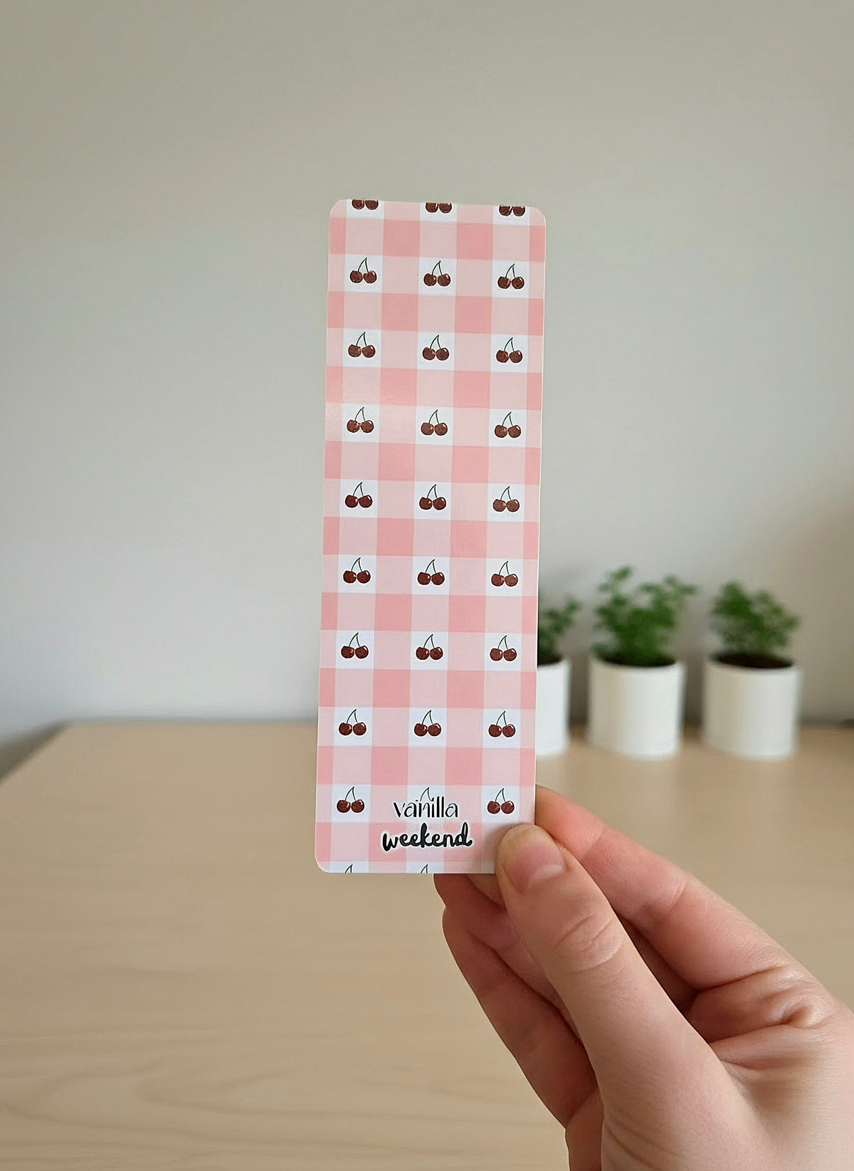 A hand holding a bookmark with a pink and white gingham pattern with mini cherries in the white boxes. The words Vanilla Weekend are at the bottom.