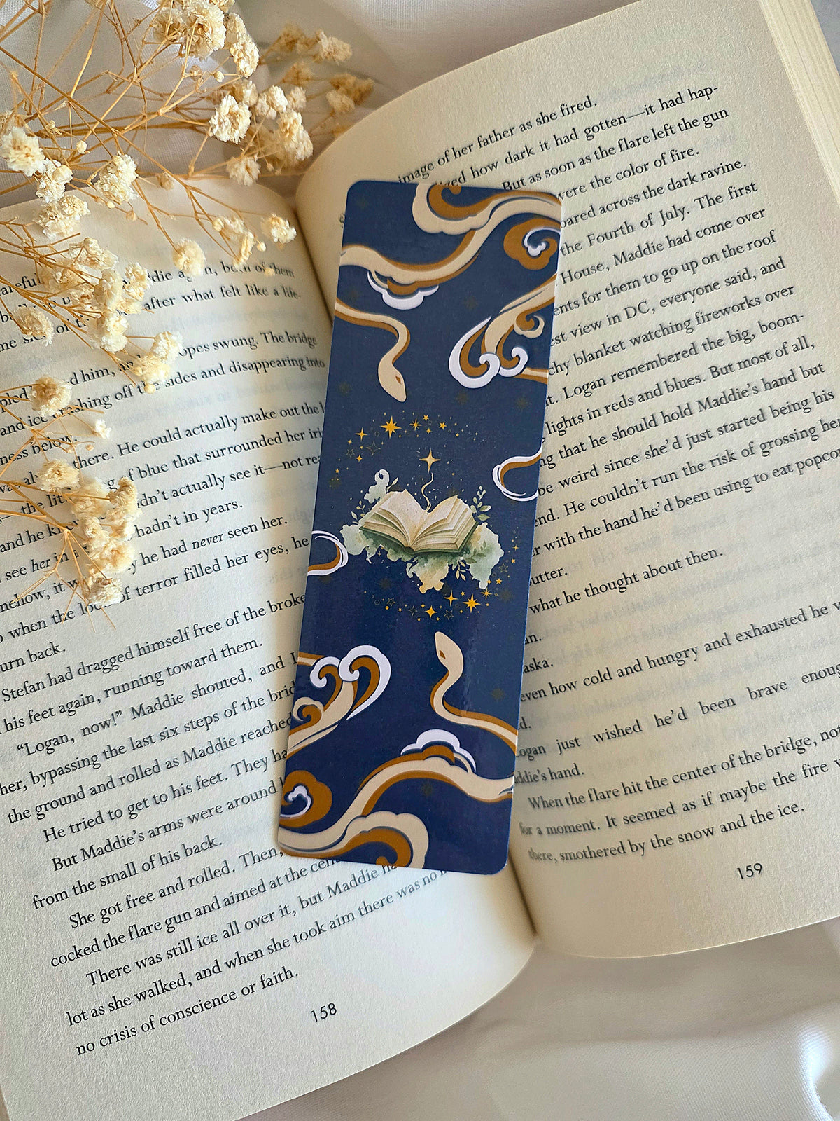 A hand holding a bookmark with a dark blue background, mysterious tan and brown snakes on the top and bottom, with a light green open book in the middle with stars swirling around. The bookmark is laying on an open book with baby's breath flowers on the left.