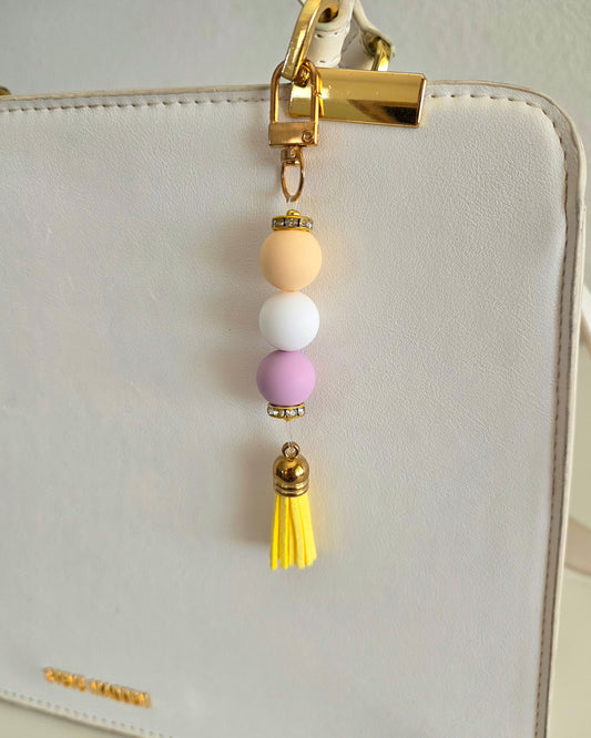 A pastel beaded keychain charm with gold-tone hardware, featuring yellow, white, and lavender beads above a yellow tassel, shown hanging from a white textured handbag.