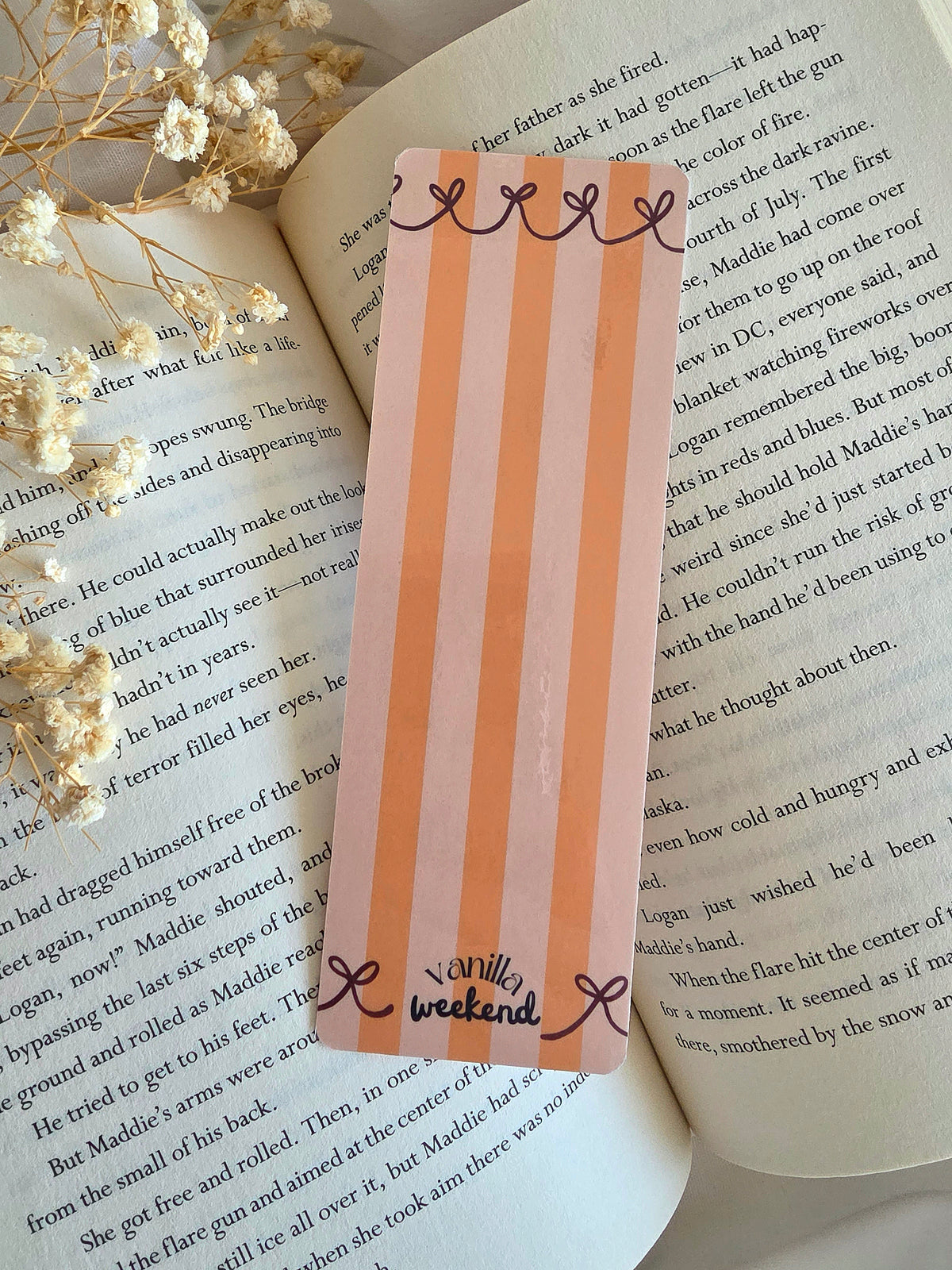 A bookmark with orange stripes on a light orange background, red ribbons lining the top and bottom, and the words Vanilla Weekend in black on the bottom. The bookmark is laying on an open book with some baby's breath flowers on the left.
