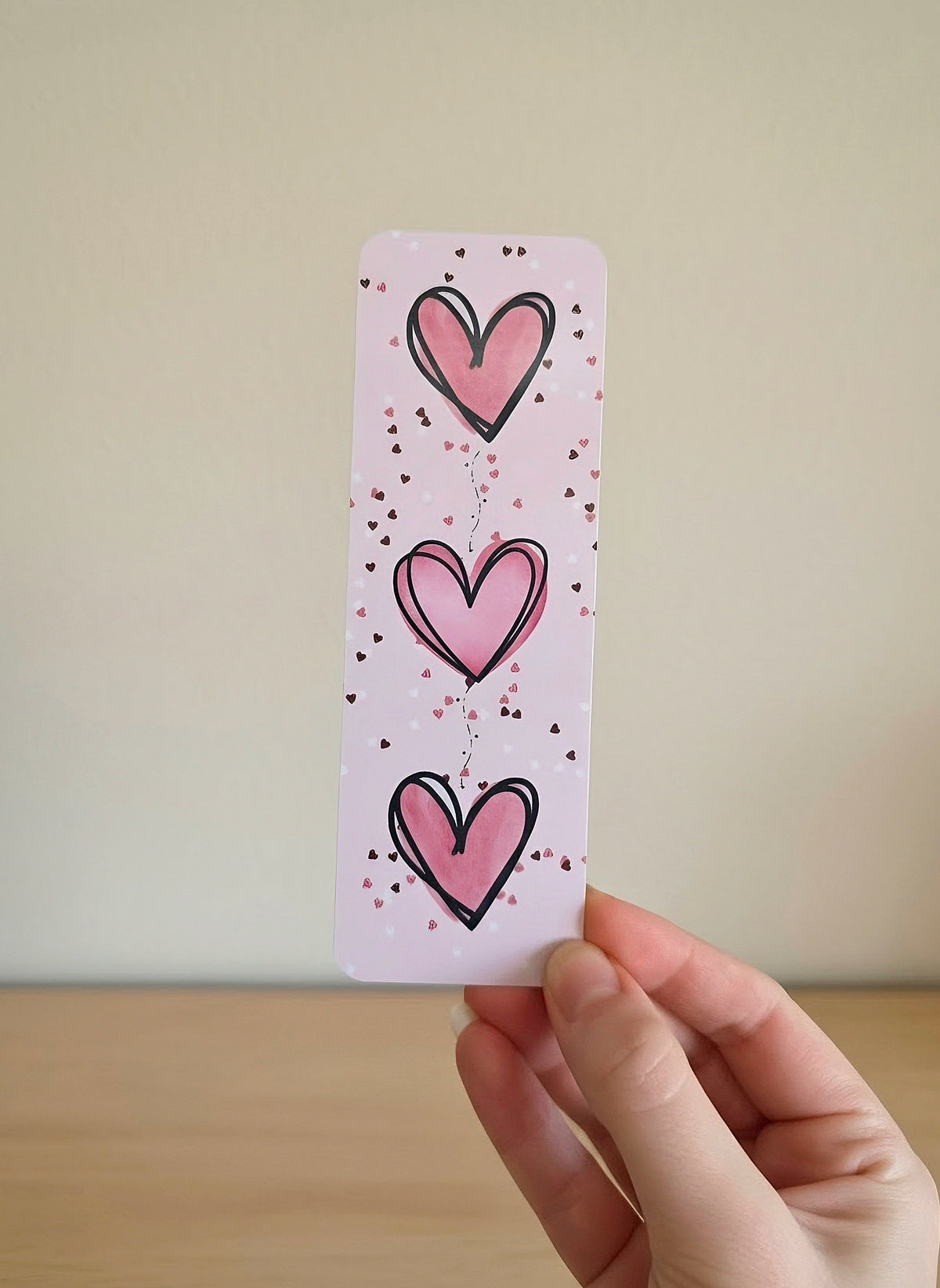A hand holding a pink bookmark with three big pink hearts and white, pink, and dark red heart confetti print.