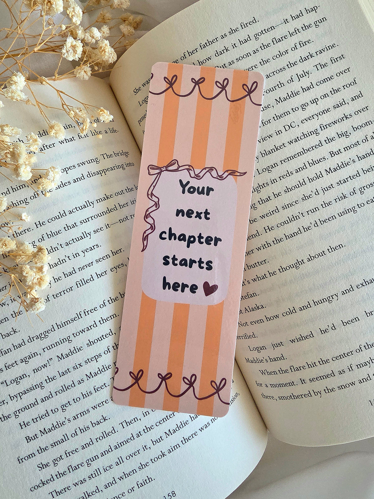 A bookmark with orange stripes on a light orange background, red ribbons lining the top and bottom, and a cream colored box in the middle with a red bow on the top left and overlay saying "Your next chapter starts here" with a dark red heart. The bookmark is laying on an open book with some baby's breath flowers on the left.