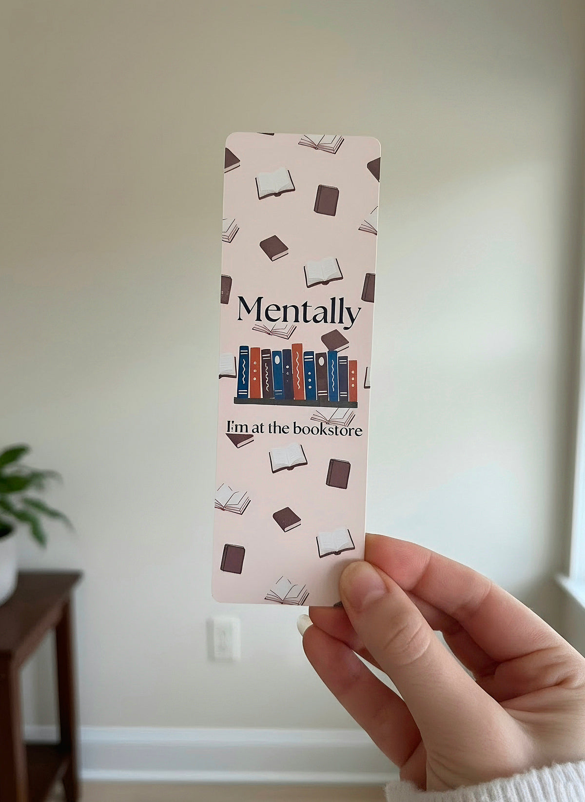 A hand holding a cream colored bookmark with books in the background. An overlay of "Mentally I'm at the bookstore" is written on the front with a shelf of blue, orange, and purple books. The bookmark is laying on an open book with some baby's breath flowers on the left.
