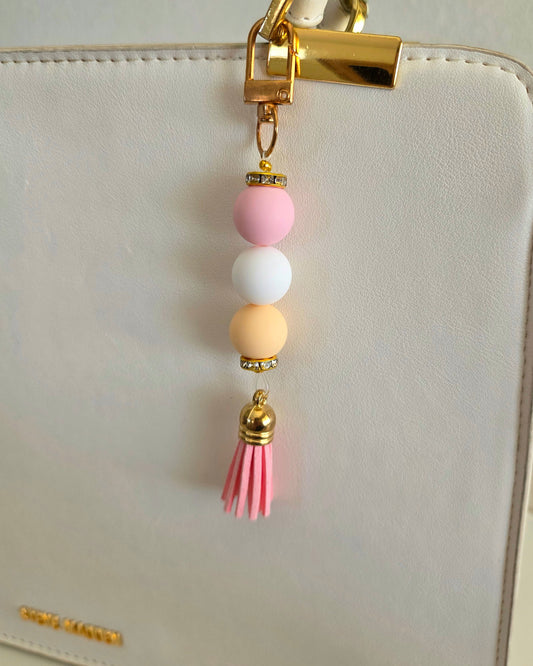 A pastel beaded keychain charm with pink, white, and yellow beads, gold-tone metal accents, and a pink tassel, shown hanging from a gold clasp on a white handbag.