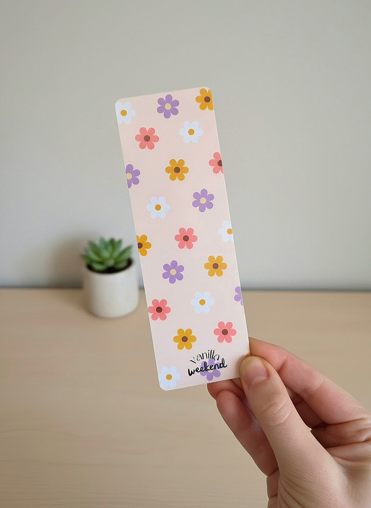 A hand holding a bookmark with simple flower pattern design with yellow, pink, white, and purple daisies on a light pink / cream background. At the bottom says Vanilla Weekend.
