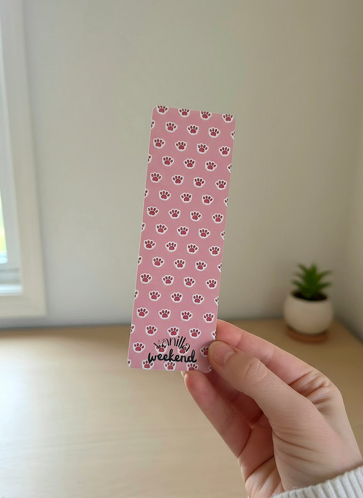 A hand holding a bookmark with a pink background and print of light pink kitty paw prints. There is a desk in the background with a potted plant.
