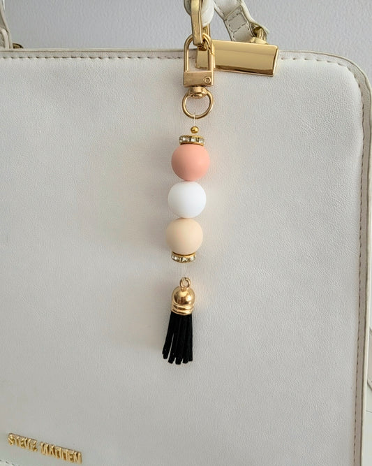 A close-up of a decorative gold-tone keychain charm with pastel pink, white, and beige beads and a black tassel clipped to a white handbag.