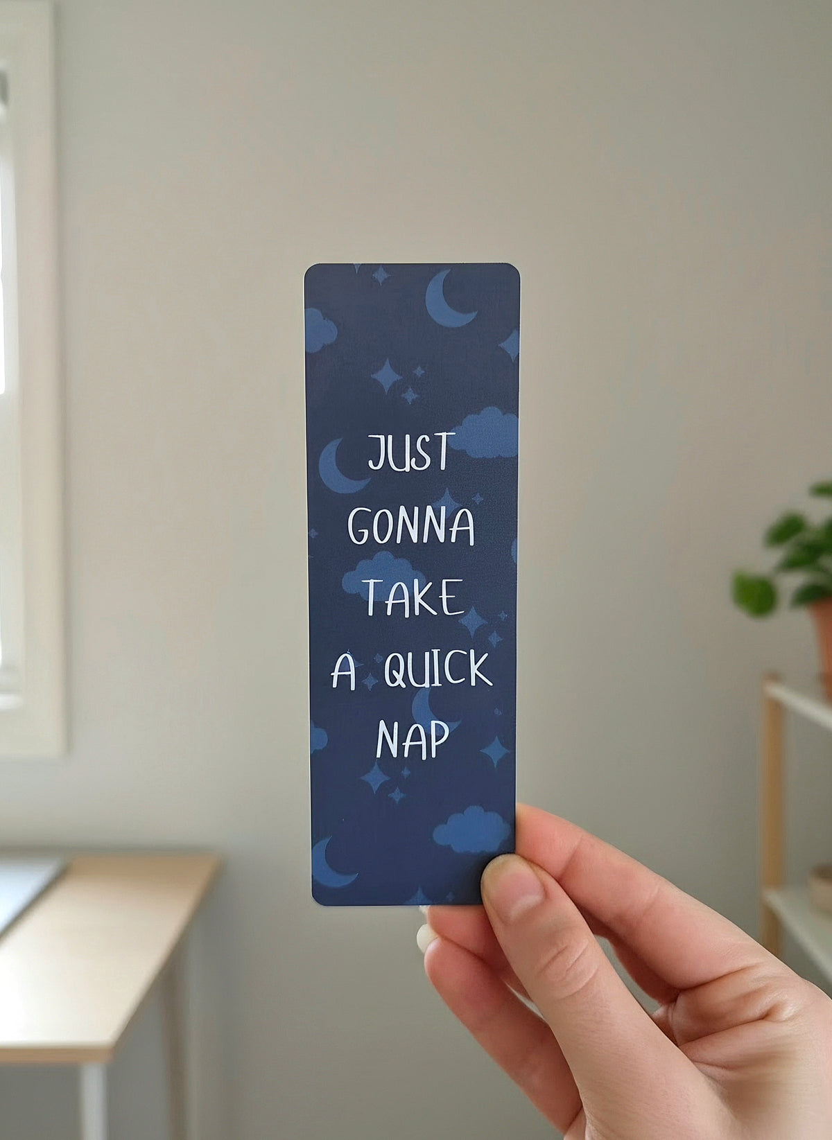 A hand holding a dark navy blue bookmark with a pattern of the moon, stars, and clouds in the background. An overlay of "Just gonna take a quick nap" is written on the bookmark in white.