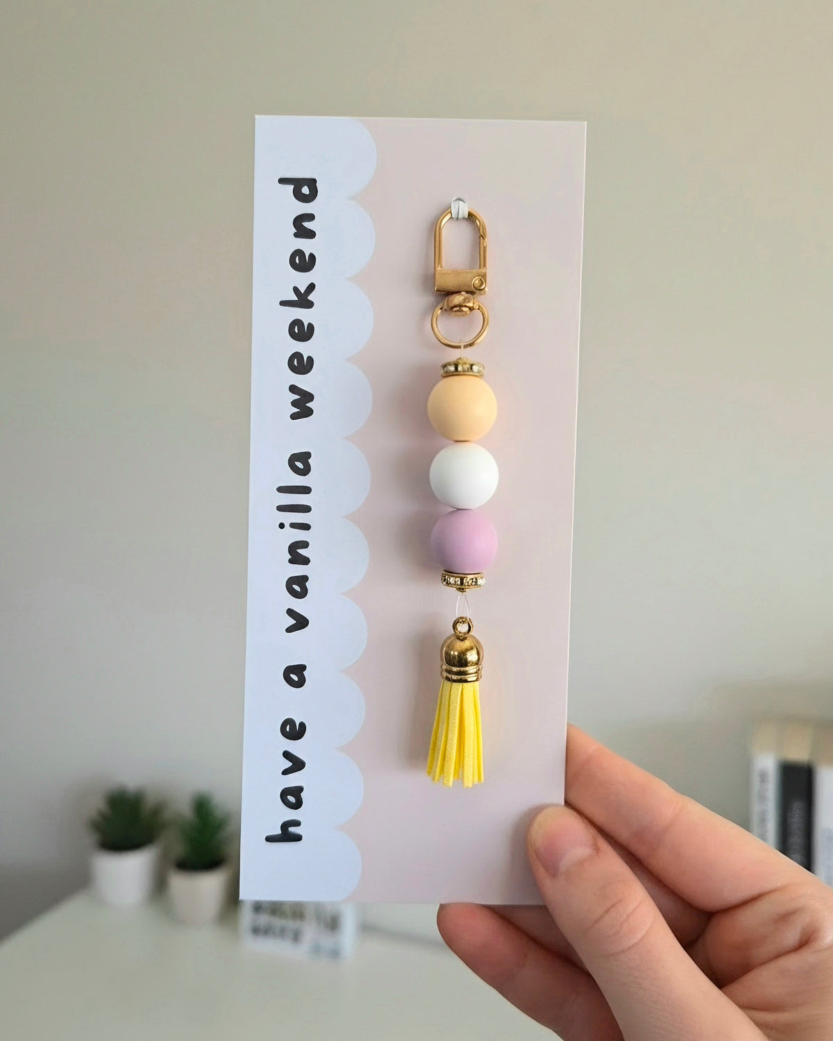 A hand holds a card featuring a pastel beaded keychain with a yellow bead, white bead, and purple bead, with gold-tone hardware, a yellow tassel, and a gold clasp. The backing card reads 'have a vanilla weekend' on a light background.