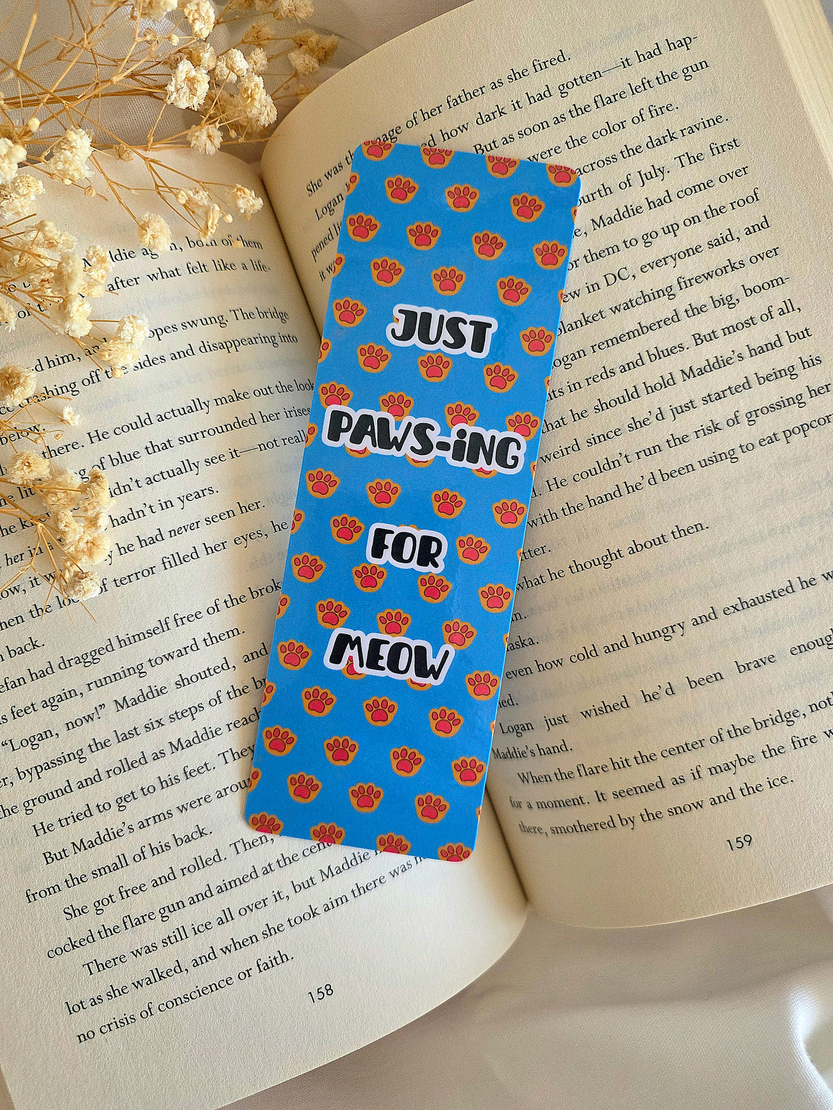 A bookmark with a blue background and orange kitty paws with overlay statement of "Just Paws-ing for Meow." Bookmark is laying on an open book with some baby's breath flowers on the left.