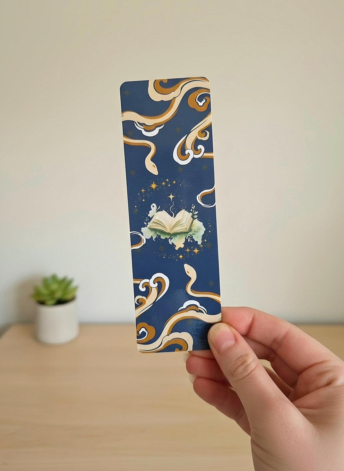 A hand holding a bookmark with a dark blue background, mysterious tan and brown snakes on the top and bottom, with a light green open book in the middle with stars swirling around.