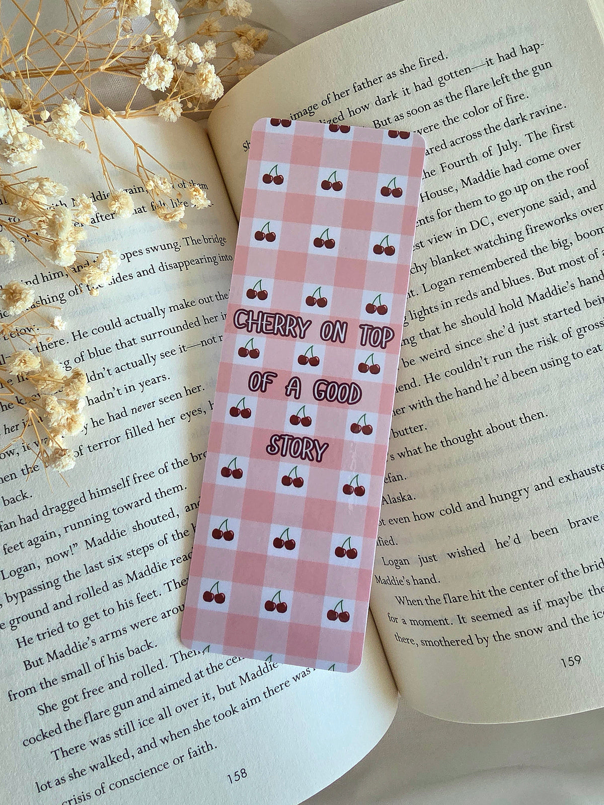 A bookmark with a pink and white gingham pattern with mini cherries in the white boxes. An overlay of "Cherry on Top of a Good Story" is written in the middle. The bookmark is laying on an open book with some baby's breath flowers on the left.