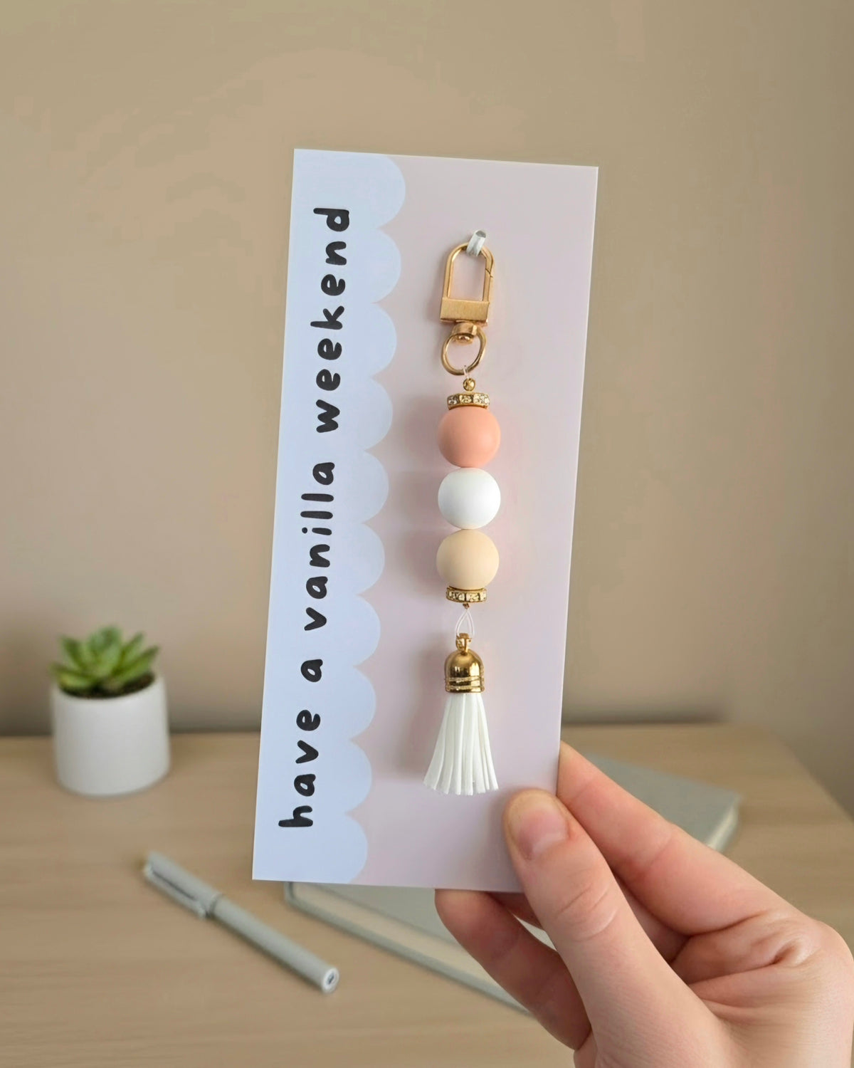 A hand holds a card featuring a decorative keychain charm with pastel peach, white, and cream beads, gold-tone lobster clasp and spacers, and a white tassel. The card reads 'have a vanilla weekend' and is photographed in a softly lit indoor setting with a small plant and desk items blurred in the background.
