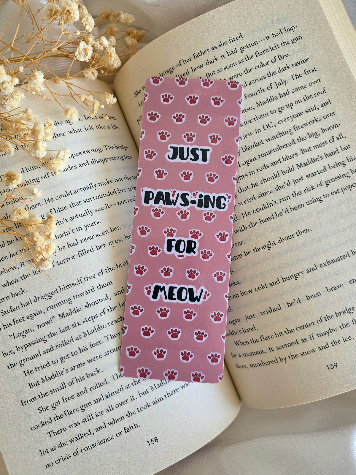 A bookmark with a pink background and light pink kitty paws with overlay statement of "Just Paws-ing for Meow." Bookmark is laying on an open book with some baby's breath flowers on the left.