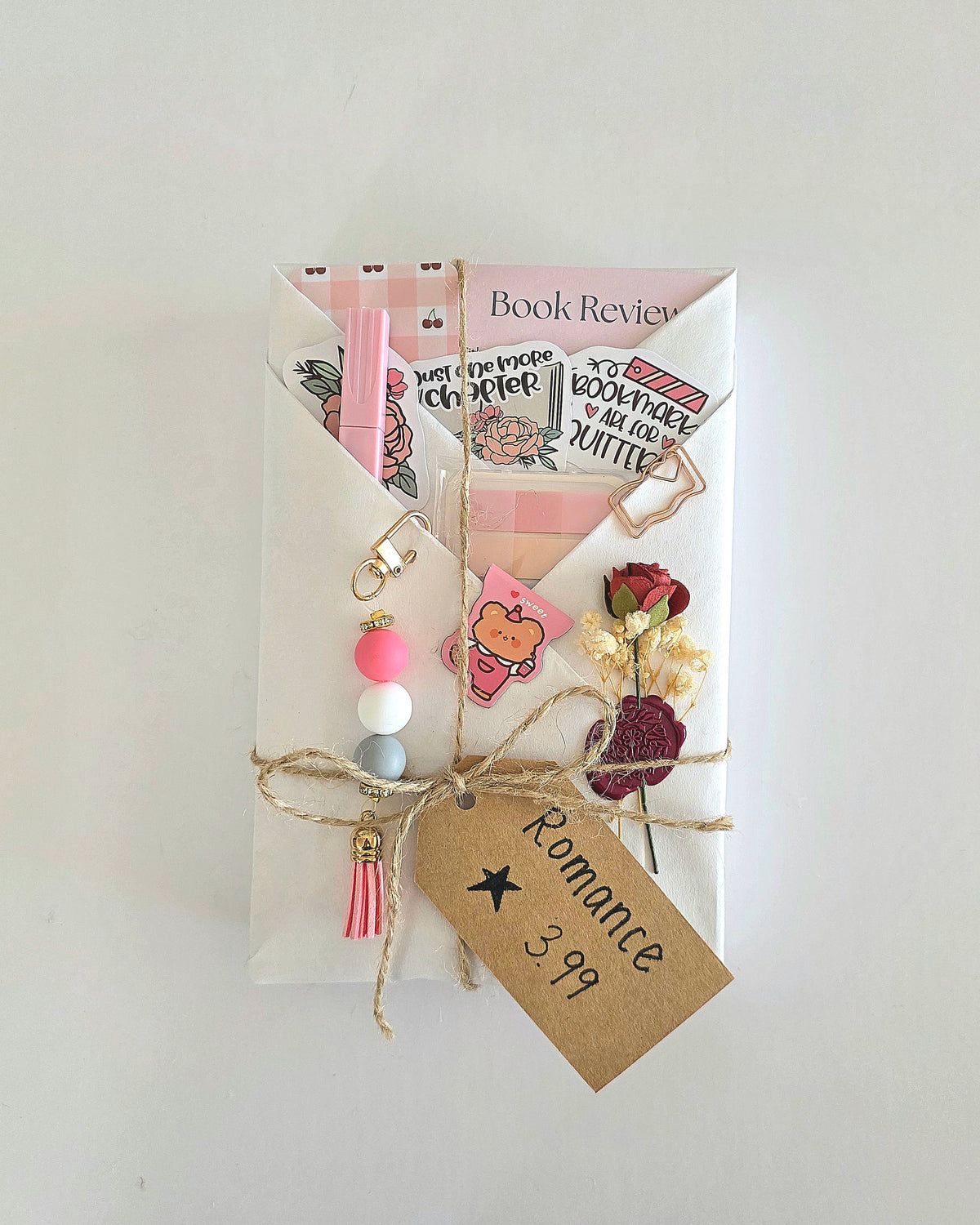 A book wrapped in white kraft paper with twine, decorated with pink elements, a small dried rose wax sealed to the wrapping, and a brown paper tag that reads 'Romance 3.99 stars.'