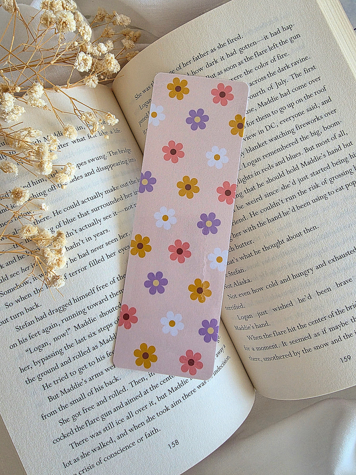 A light pink / cream bookmark with simple flower pattern design with yellow, pink, white, and purple daisies. The bookmark is laying on an open book with baby's breath flowers on the left.