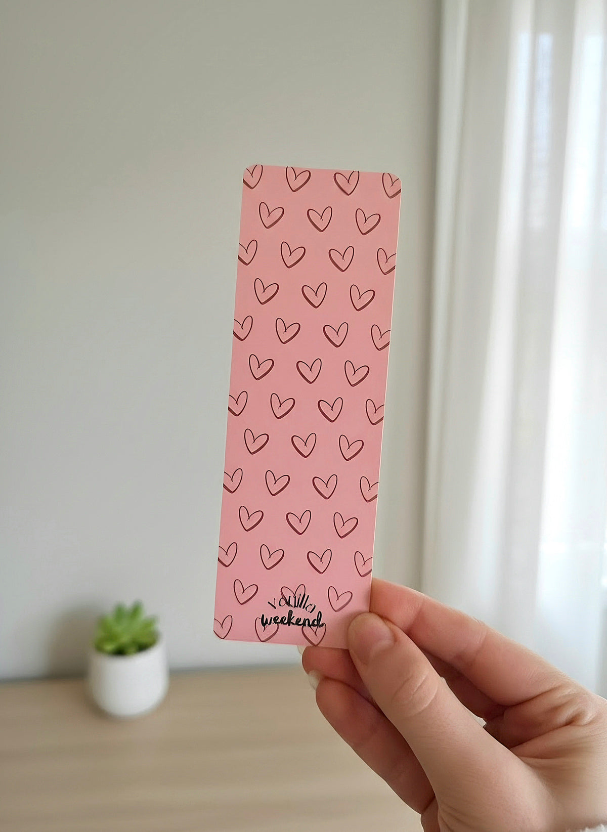 A hand holding a pink bookmark with a pattern of outlined hearts in a dark red color. 