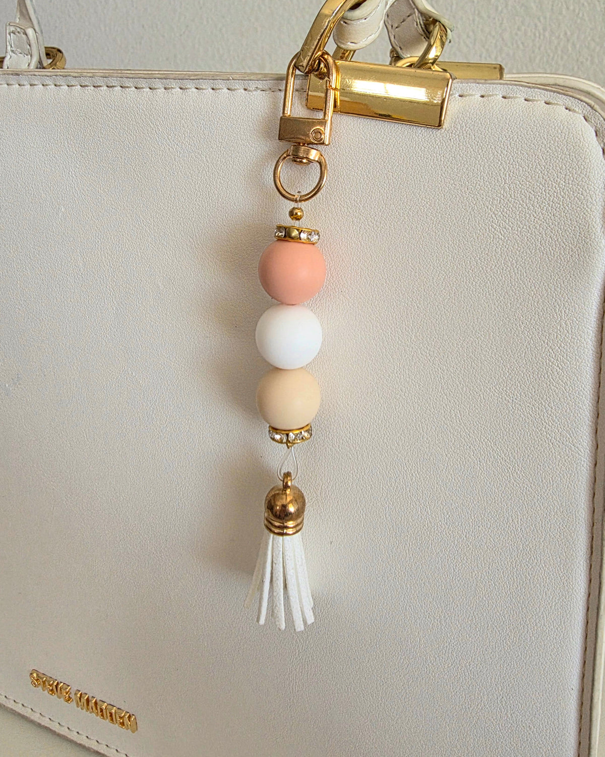 A pastel beaded keychain charm with gold-tone lobster clasp and spacers, featuring peach, white, and beige beads and a white tassel, shown hanging from a white handbag.