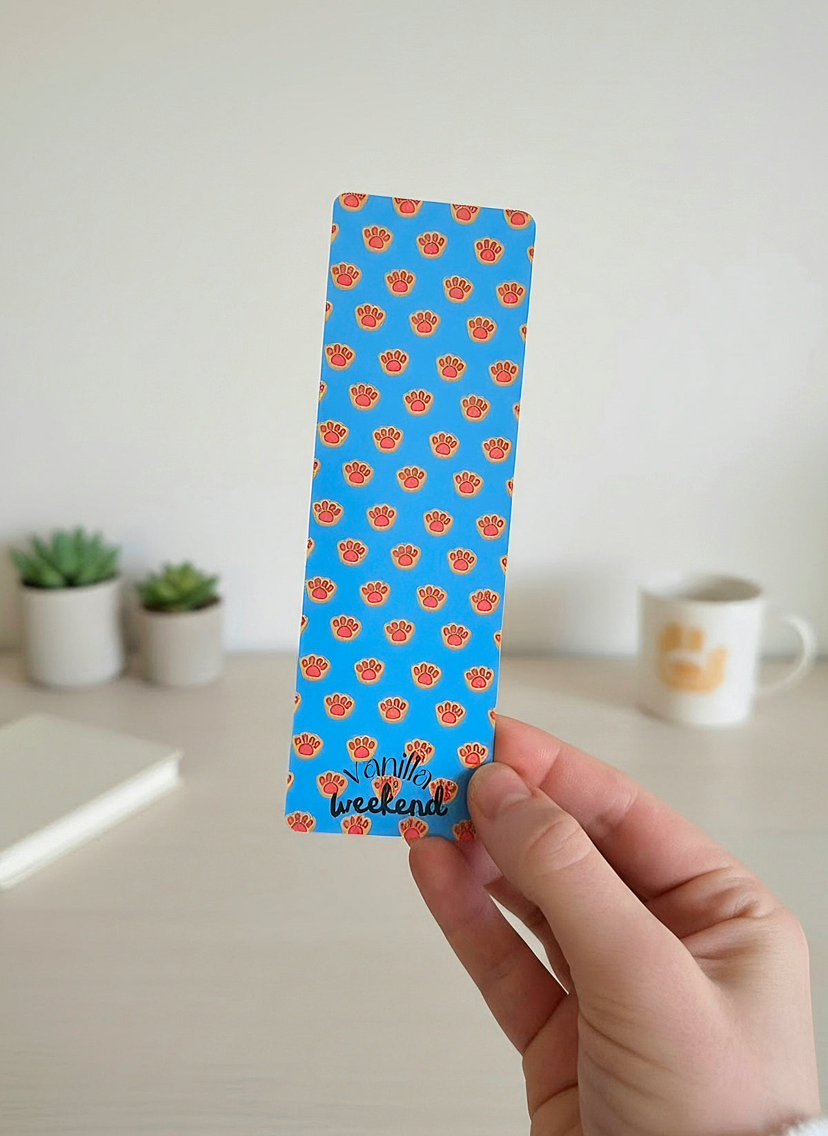 A hand holding a bookmark with a blue background and print of orange kitty paw prints. There is a desk in the background with a cup, potted plants, and a book.