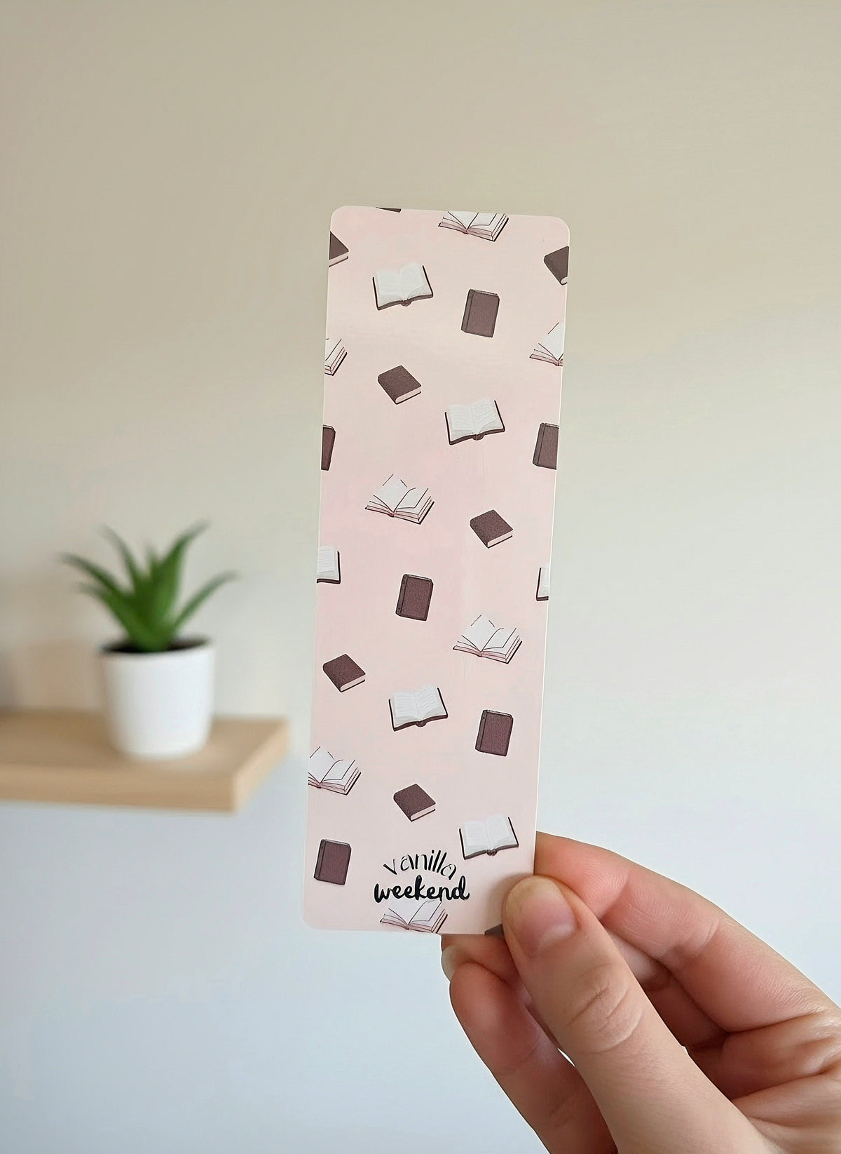 A hand holding a cream colored bookmark with books in the background. The words Vanilla Weekend are at the bottom.