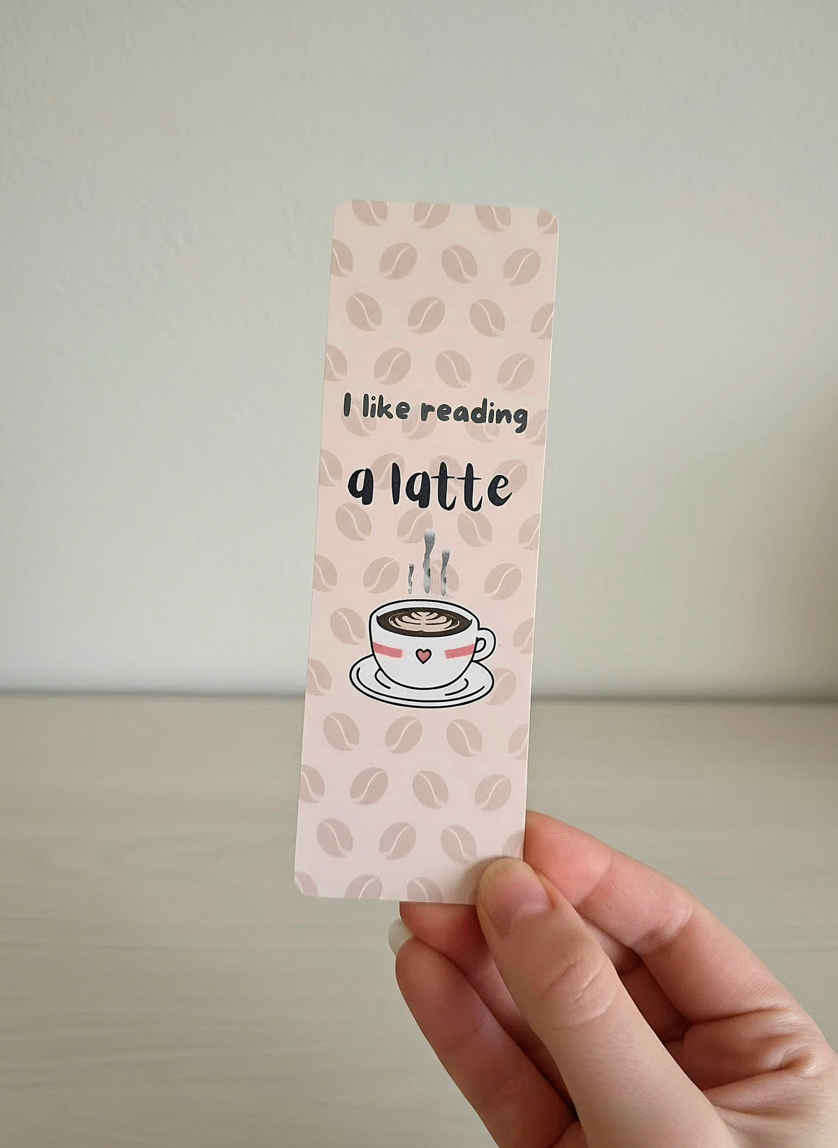 A hand holding a cream colored bookmark with a faded coffee bean pattern in the background. An overlay of "I like reading a latte" and a steamy cup of coffee is on the front. 