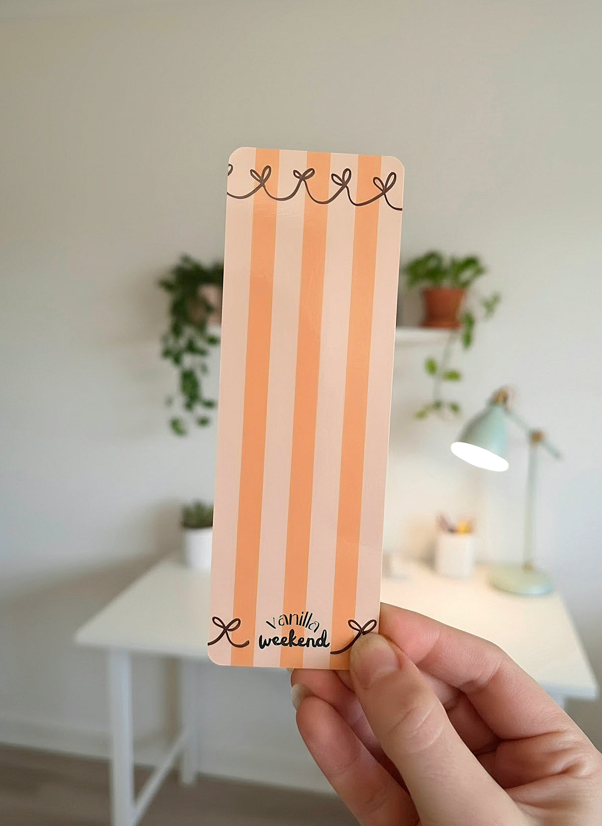 A hand holding a bookmark with orange stripes on a light orange background, red ribbons lining the top and bottom, and the words Vanilla Weekend in black on the bottom.