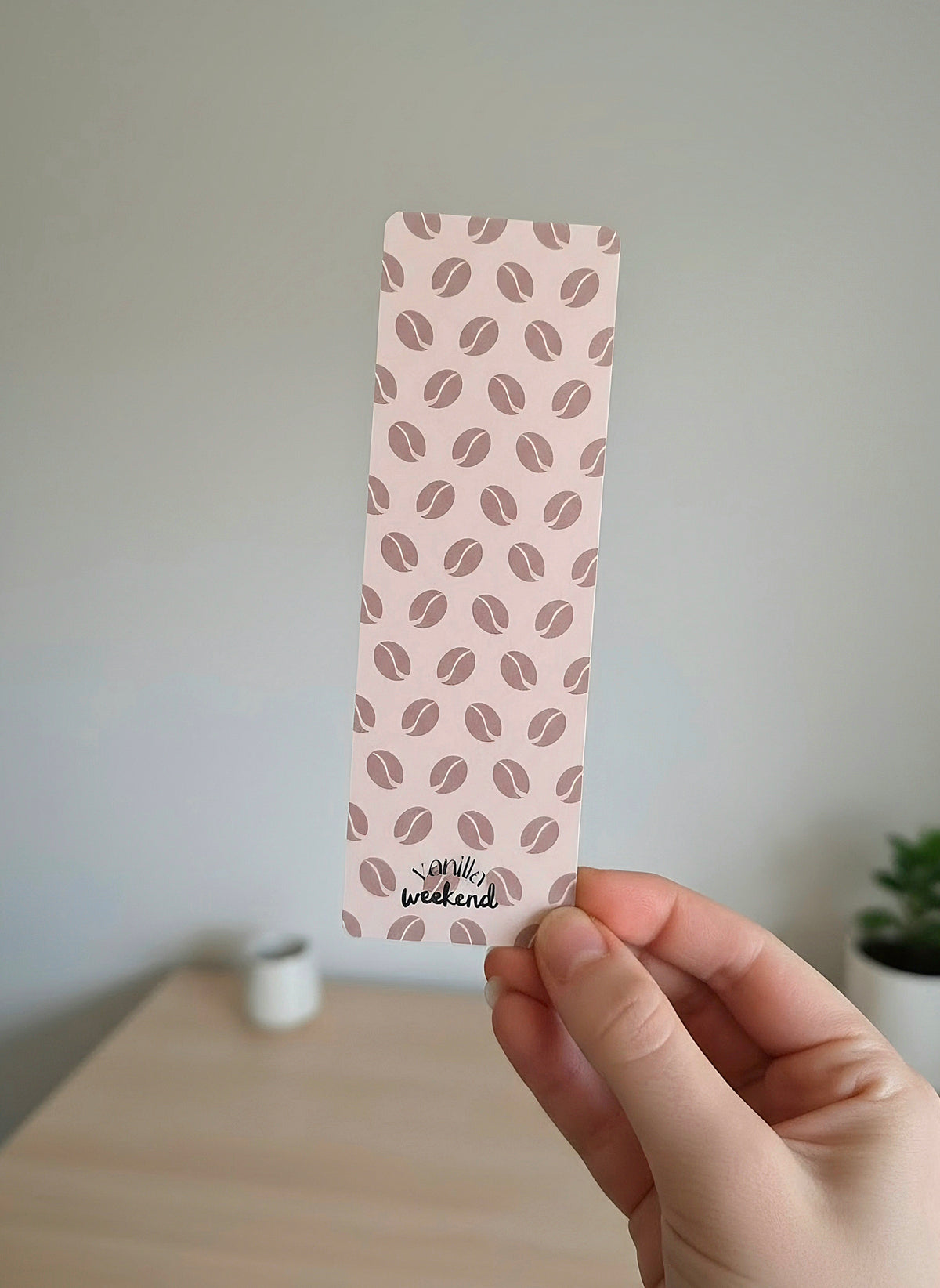 A hand holding a cream colored bookmark with a coffee bean pattern in the background. The words Vanilla Weekend are at the bottom. 