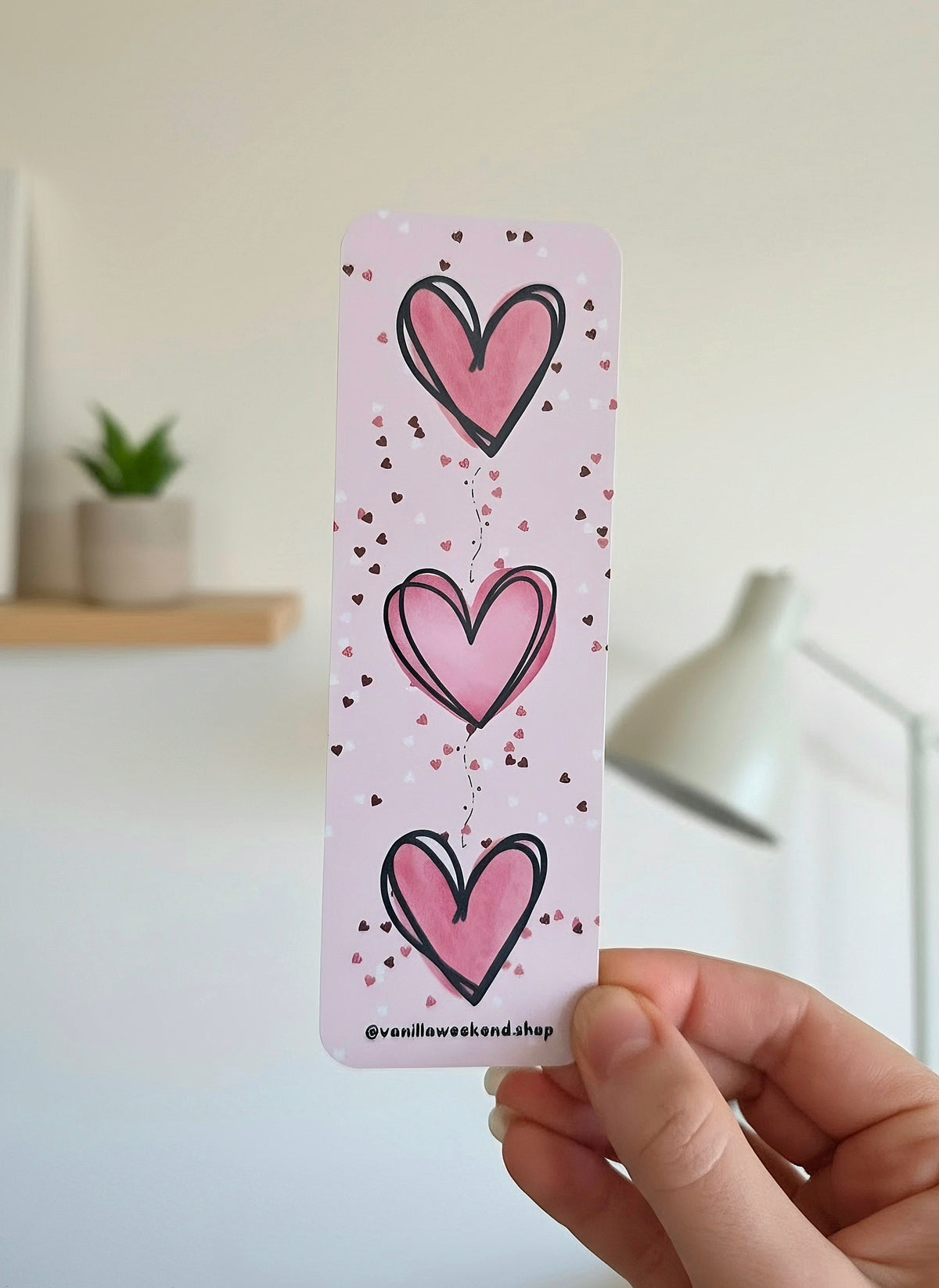 A hand holding a pink bookmark with three big pink hearts and white, pink, and dark red heart confetti print with @vanillaweekend.shop in black at the bottom.