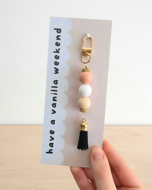 A hand holding a card featuring a decorative keychain or bag charm with gold-tone lobster clasp and spacers, pastel peach, white, and cream beads, and a black tassel. The card reads 'have a vanilla weekend' on a light background.