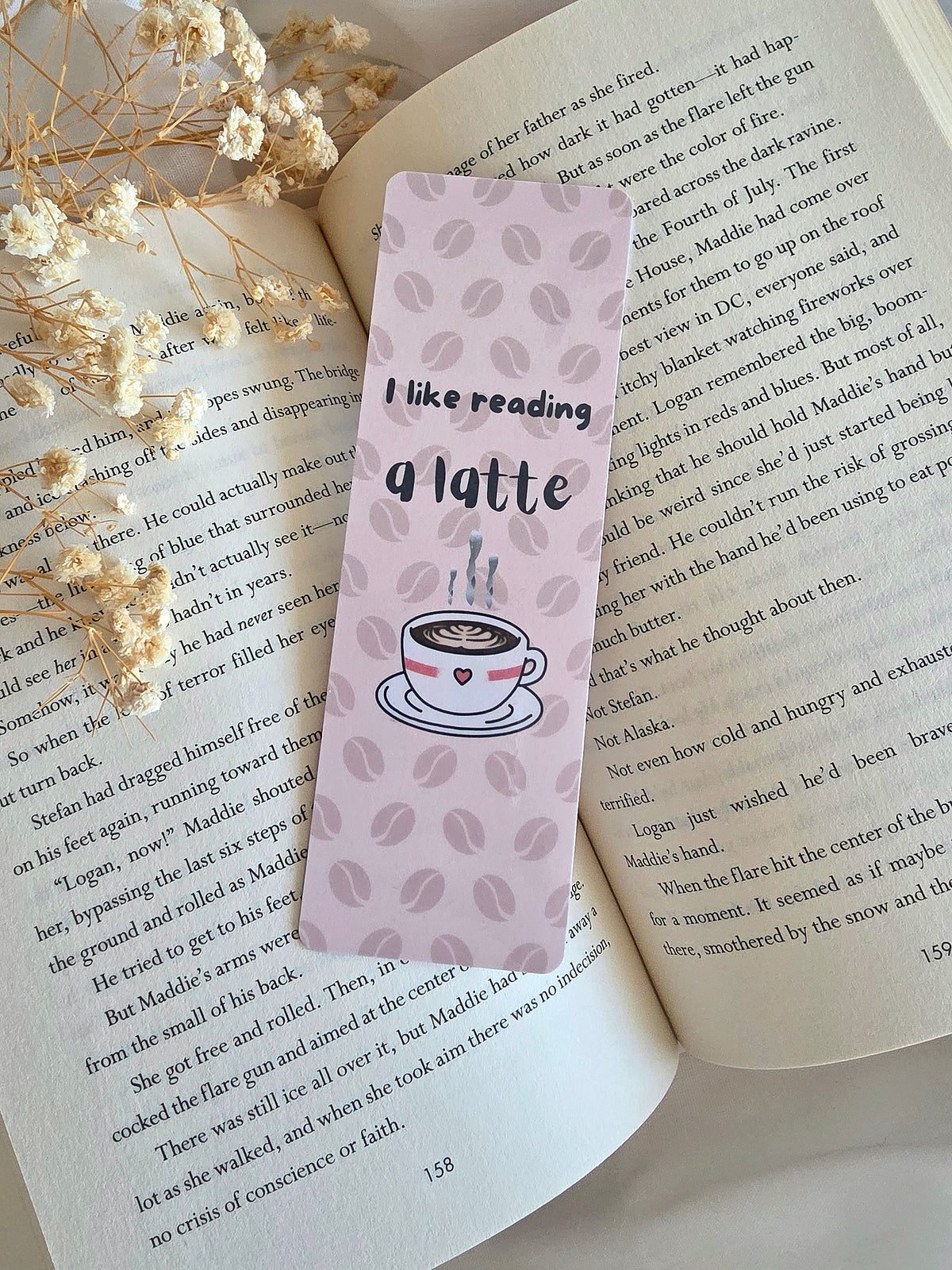 A cream colored bookmark with a faded coffee bean pattern in the background. An overlay of "I like reading a latte" and a steamy cup of coffee is on the front. The bookmark is laying on an open book with some baby's breath flowers on the left.