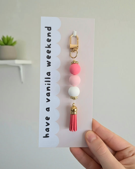 A hand holding a white card featuring a decorative keychain charm. The charm has a gold-tone clasp, pink and light pink beads, a white bead, and a pink tassel.