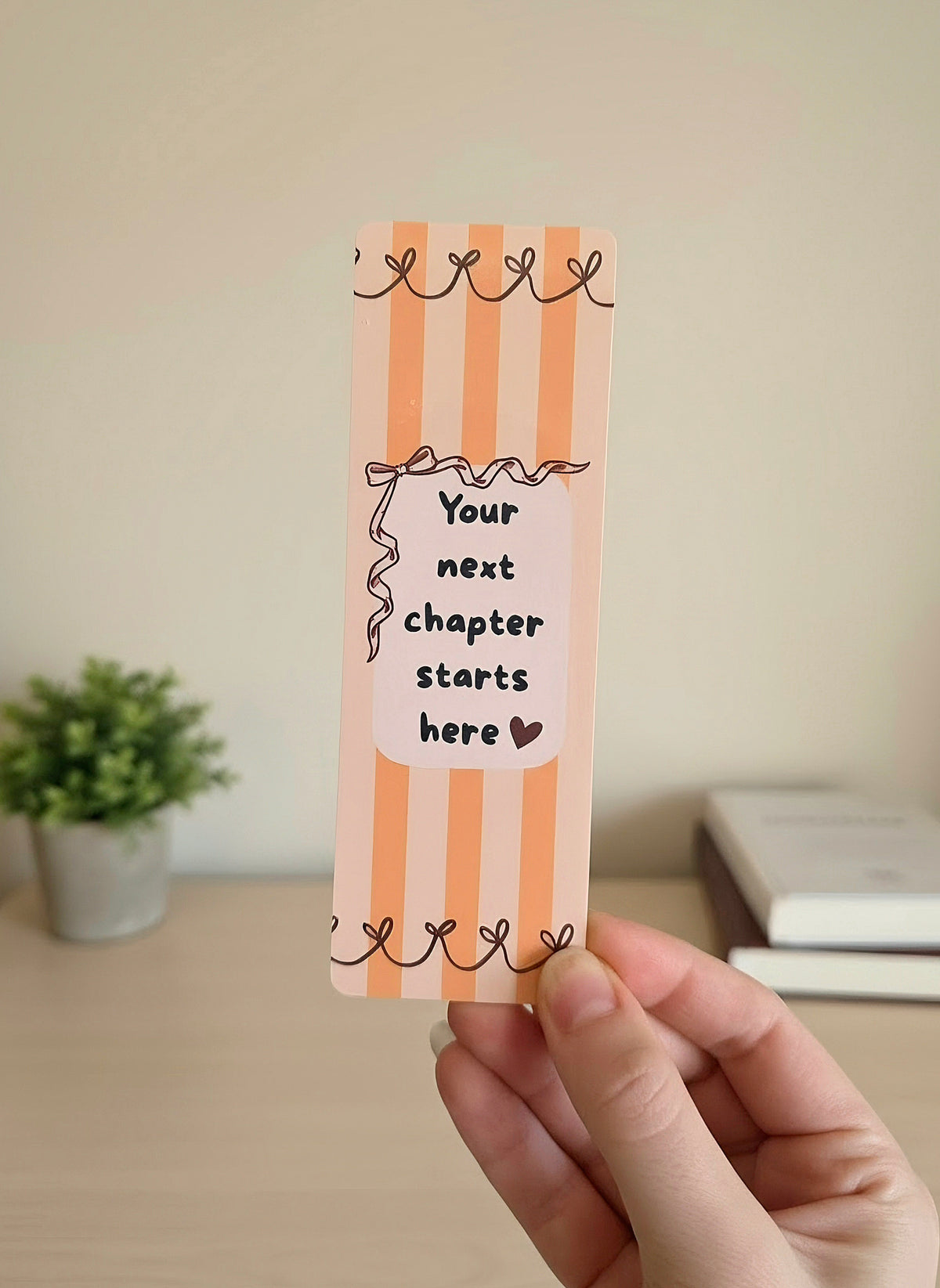 A hand holding a bookmark with orange stripes on a light orange background, red ribbons lining the top and bottom, and a cream colored box in the middle with a red bow on the top left and overlay saying "Your next chapter starts here" with a dark red heart. 