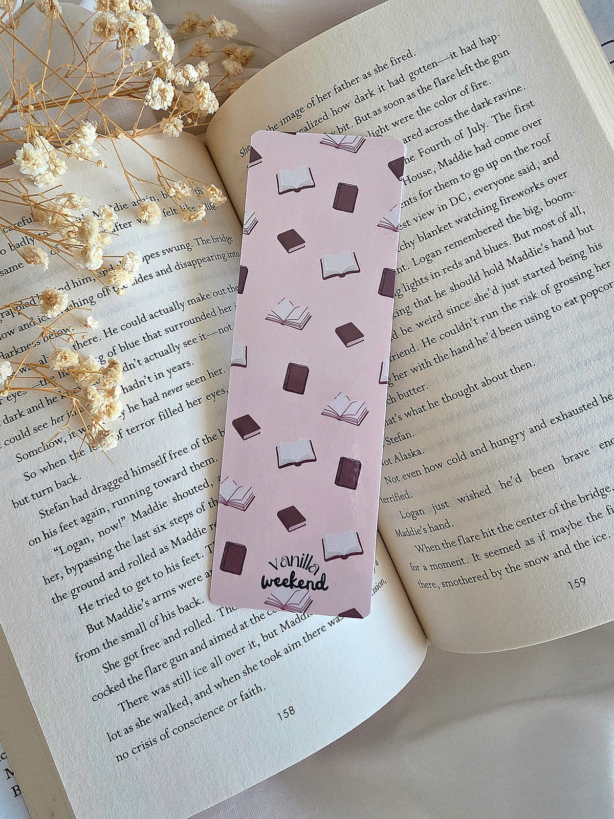 A cream colored bookmark with books in the background. The words Vanilla Weekend are at the bottom. The bookmark is laying on an open book with some baby's breath flowers on the left.