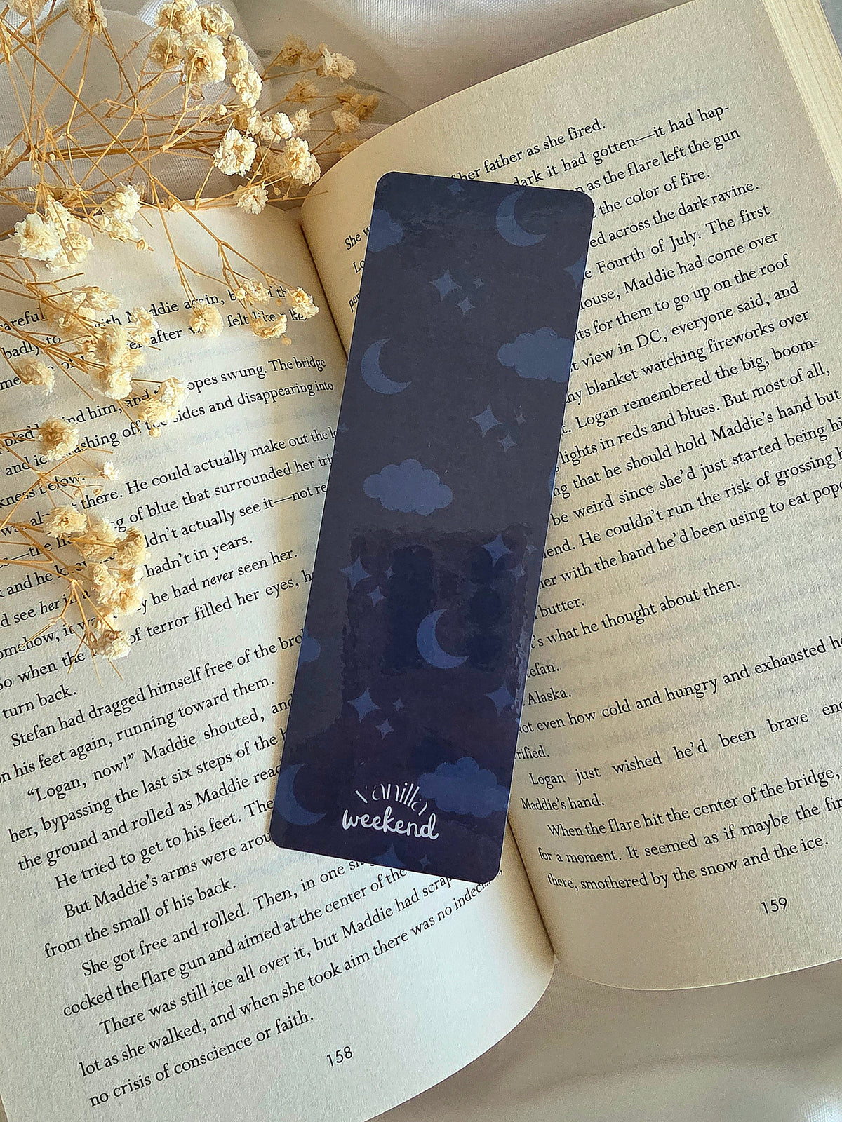 A dark navy blue bookmark with a pattern of the moon, stars, and clouds in the background. The words Vanilla Weekend are in the bottom in white. The bookmark is laying on an open book with some baby's breath flowers on the left.