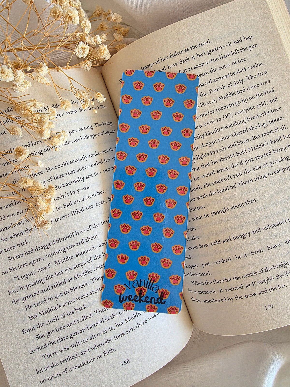 A bookmark with a blue background and orange kitty paw pattern. The bottom says Vanilla Weekend Bookmark is laying on an open book with some baby's breath flowers on the left. 
