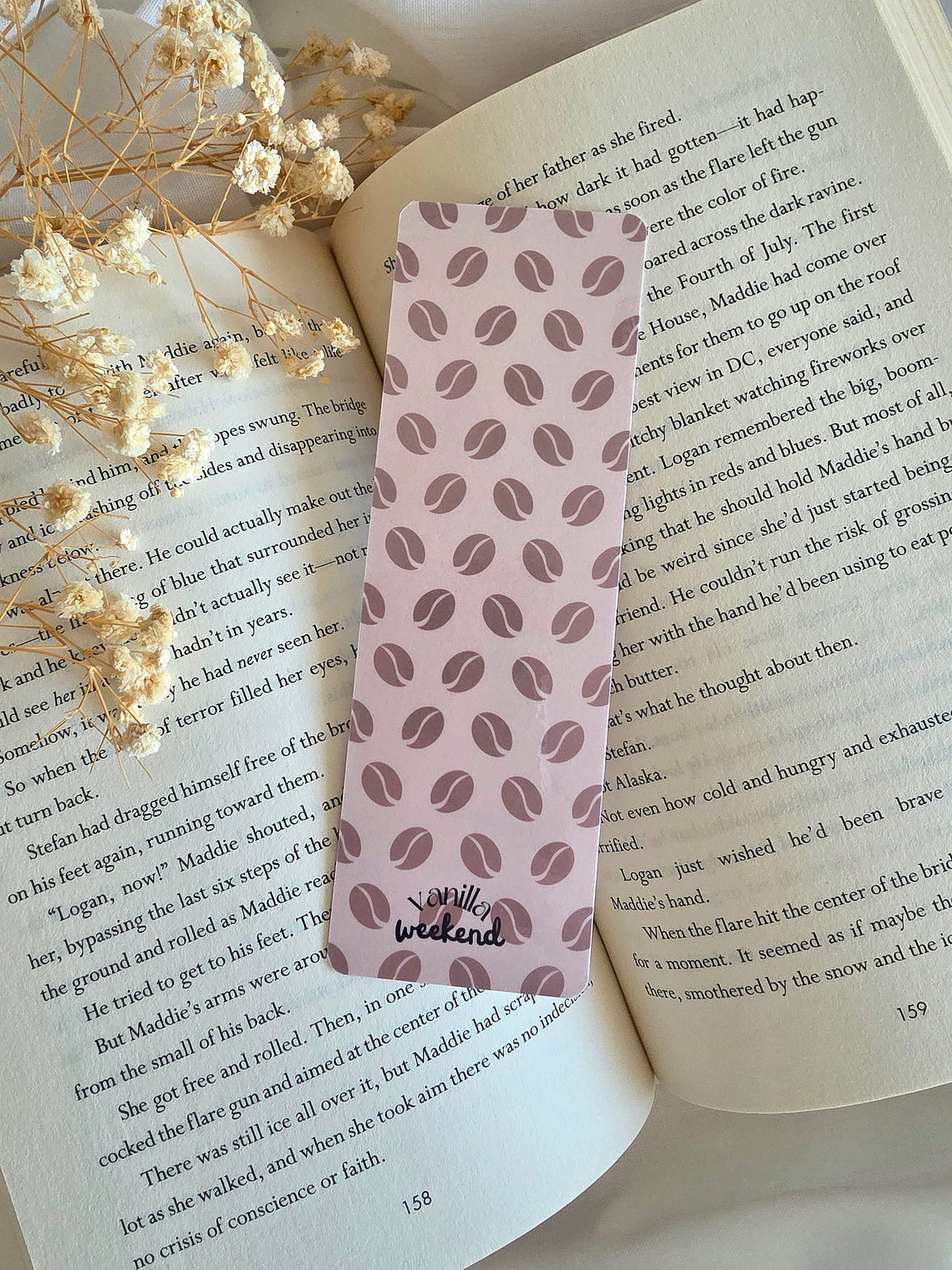 A cream colored bookmark with a coffee bean pattern in the background. The words Vanilla Weekend are at the bottom. The bookmark is laying on an open book with some baby's breath flowers on the left.