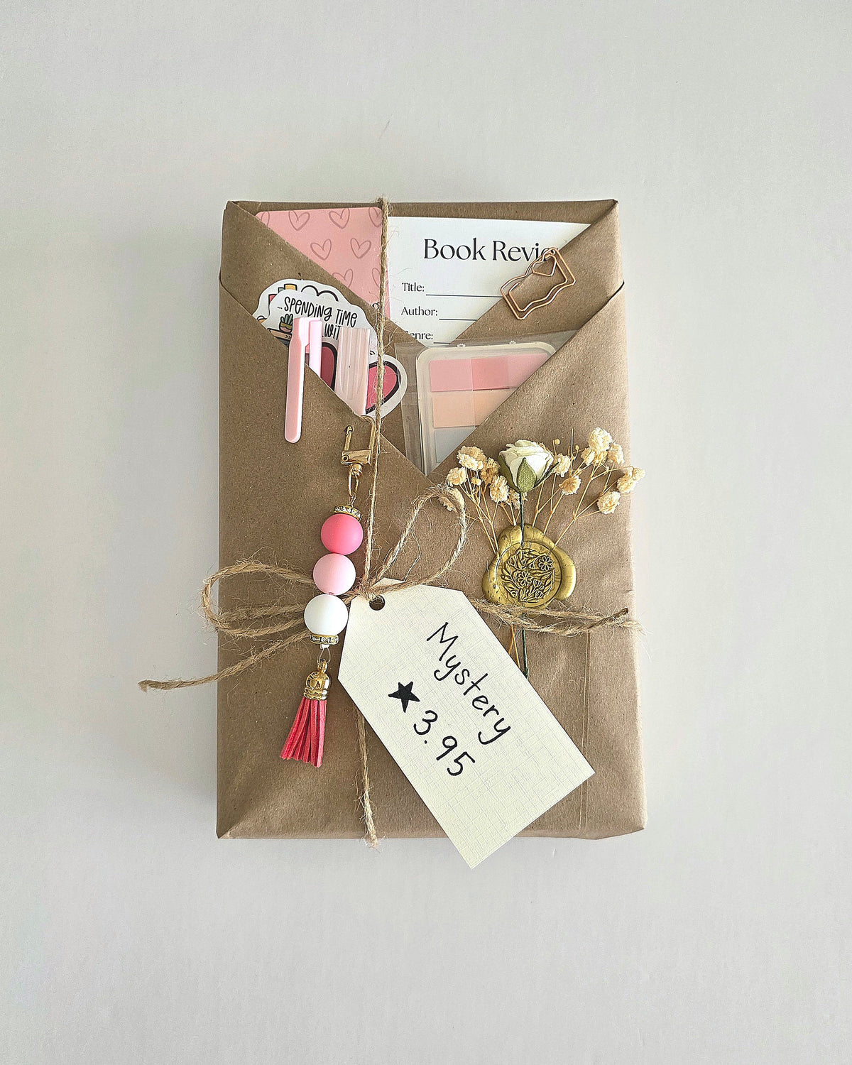 Blind date with a book - book wrapped in brown craft paper with twine and a white tag that states "Mystery, 3.95 stars." Also a white flower embellishment that is wax-stamped on the brown craft paper. There is a pink bookmark, book review card, 3 stickers, sticky notes for annotating, a pen, and a highlighter in the folds of the wrapped book. Lastly, a pink bead and tassel keychain is hanging from one of the folds. 