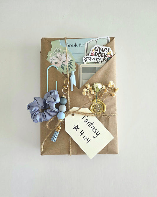 Blind date with a book - book wrapped in brown craft paper with twine and a white tag that states "Fantasy, 4.04 stars." Also a flower embellishment with baby's breath and white rose that is wax-stamped on the brown craft paper. There is a magnetic flower bookmark, book review card, 3 stickers, sticky notes for annotating, a pen, and a hair tie entwined in a large paperclip in the folds. Lastly, a blue bead and tassel keychain is hanging from one of the folds.