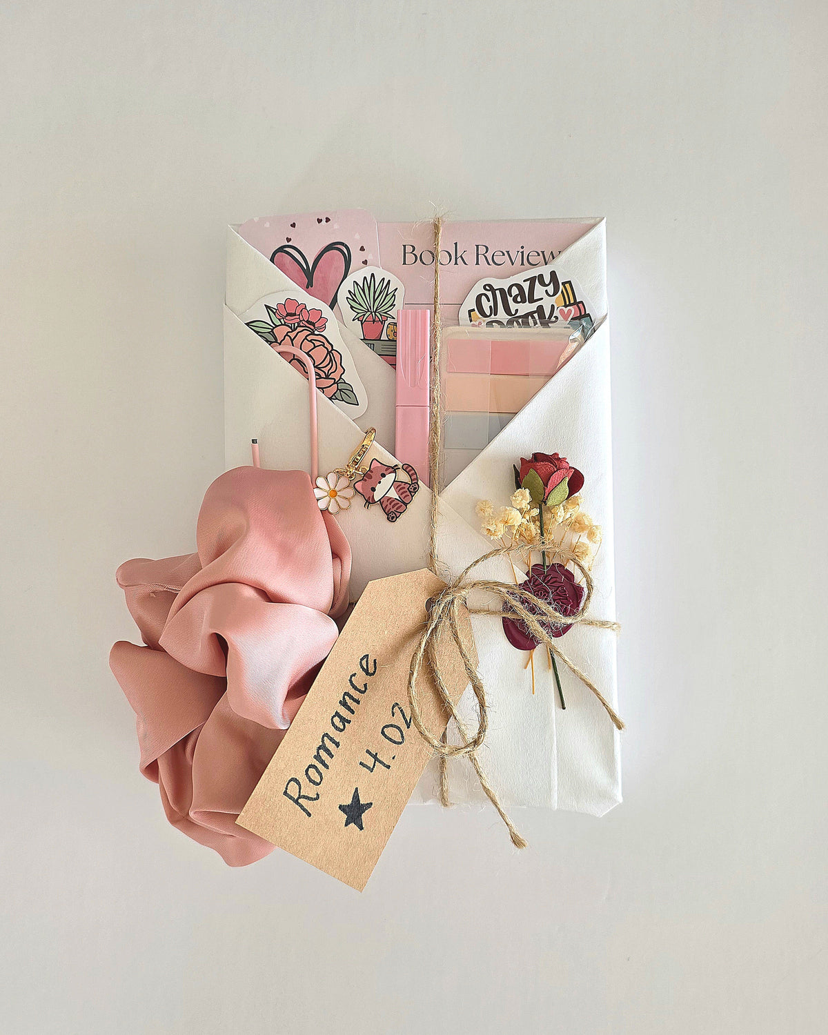 A wrapped book gift set with white wrapping paper, decorative stickers, a pink satin hair tie, and a tag reading 'Romance 4.02' against a plain light background. Includes a cat and daisy keychain and wax sealed red rose and baby's breath flower embellishments.