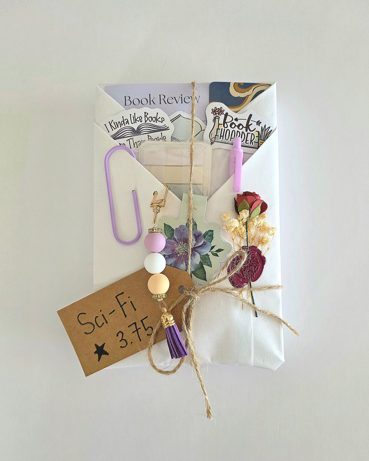 A wrapped book gift set tied with twine and decorated with dried flowers, a purple paperclip, beads, and a handwritten price tag reading 'Sci-Fi 3.75'. Has a wax-sealed flower embellishment with baby's breath and red rose.