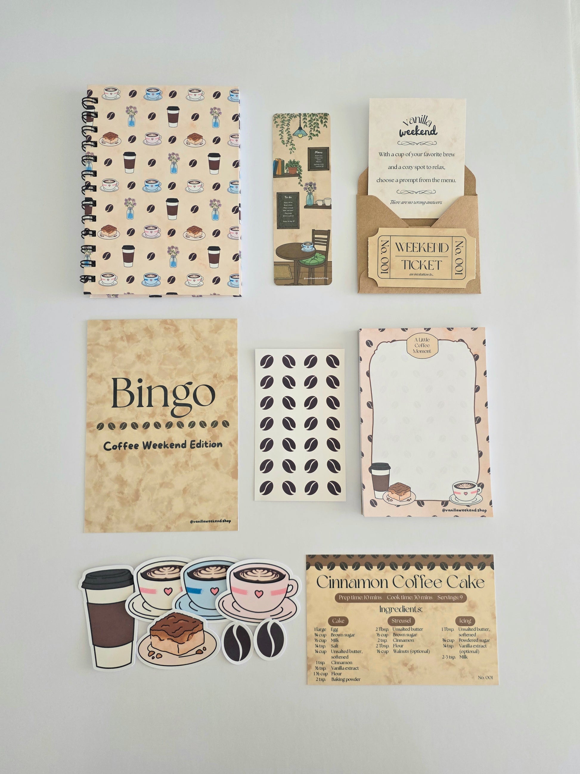A flat lay of a coffee-themed stationery set on a white background. From top left to bottom right: a spiral notebook, double sided bookmark, envelope with journal prompts and collectible weekend ticket, bingo card, coffee bean sticker sheet, notepad, sticker pack, and recipe card.
