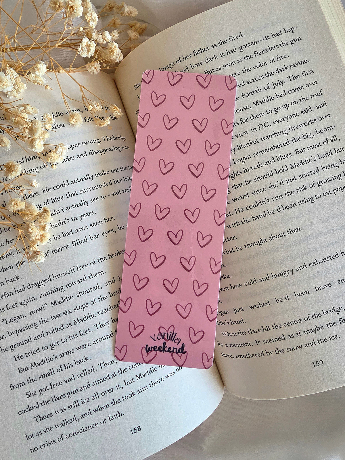 A pink bookmark with a heart pattern. The bookmark is laying on an open book with some baby's breath flowers on the left.