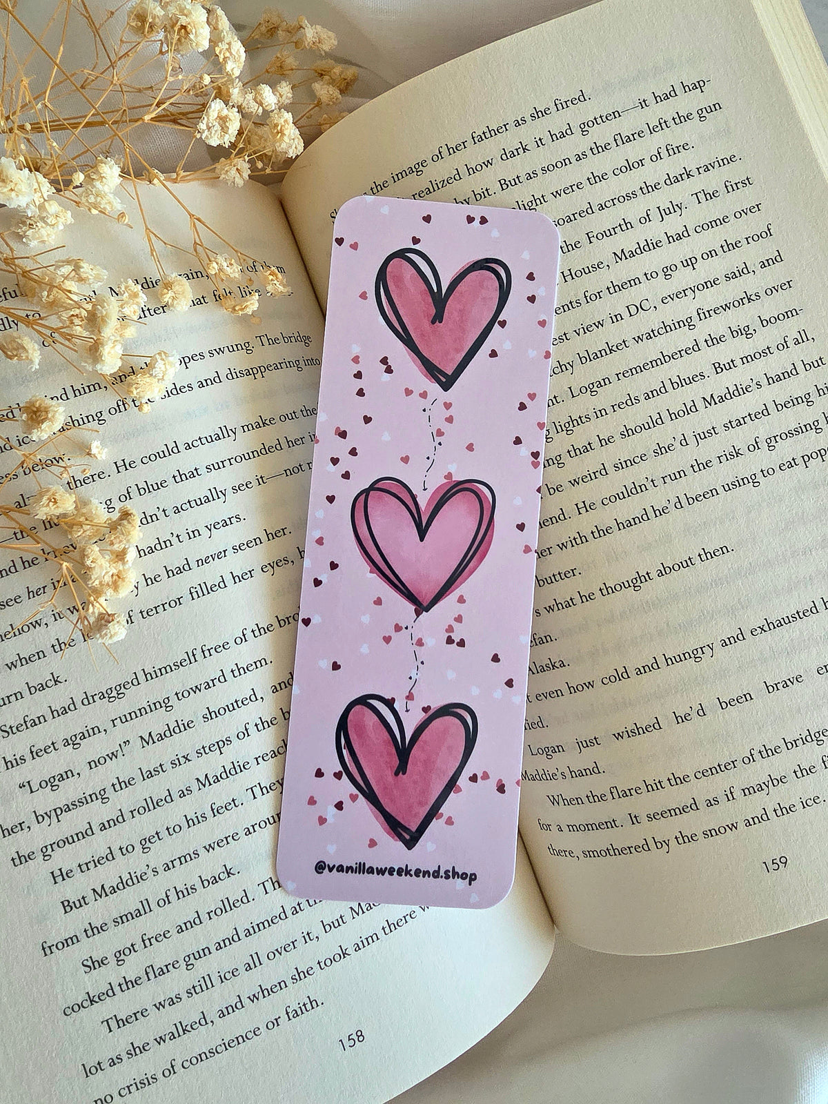 A pink bookmark with three big pink hearts and white, pink, and dark red heart confetti print with @vanillaweekend.shop in black at the bottom. The bookmark is laying on an open book with some baby's breath flowers on the left.