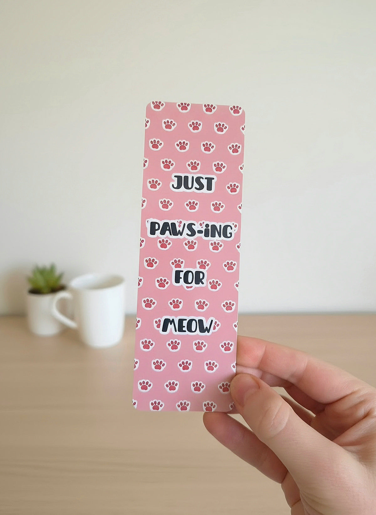 A bookmark with a pink background and light pink kitty paws with overlay statement of "Just Paws-ing for Meow." Bookmark is held by a hand with a cup and plant on a desk in the background.