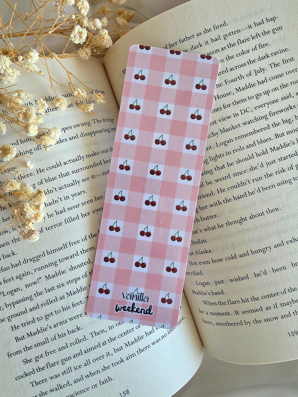 A bookmark with a pink and white gingham pattern with mini cherries in the white boxes. The words Vanilla Weekend are at the bottom. The bookmark is laying on an open book with some baby's breath flowers on the left.