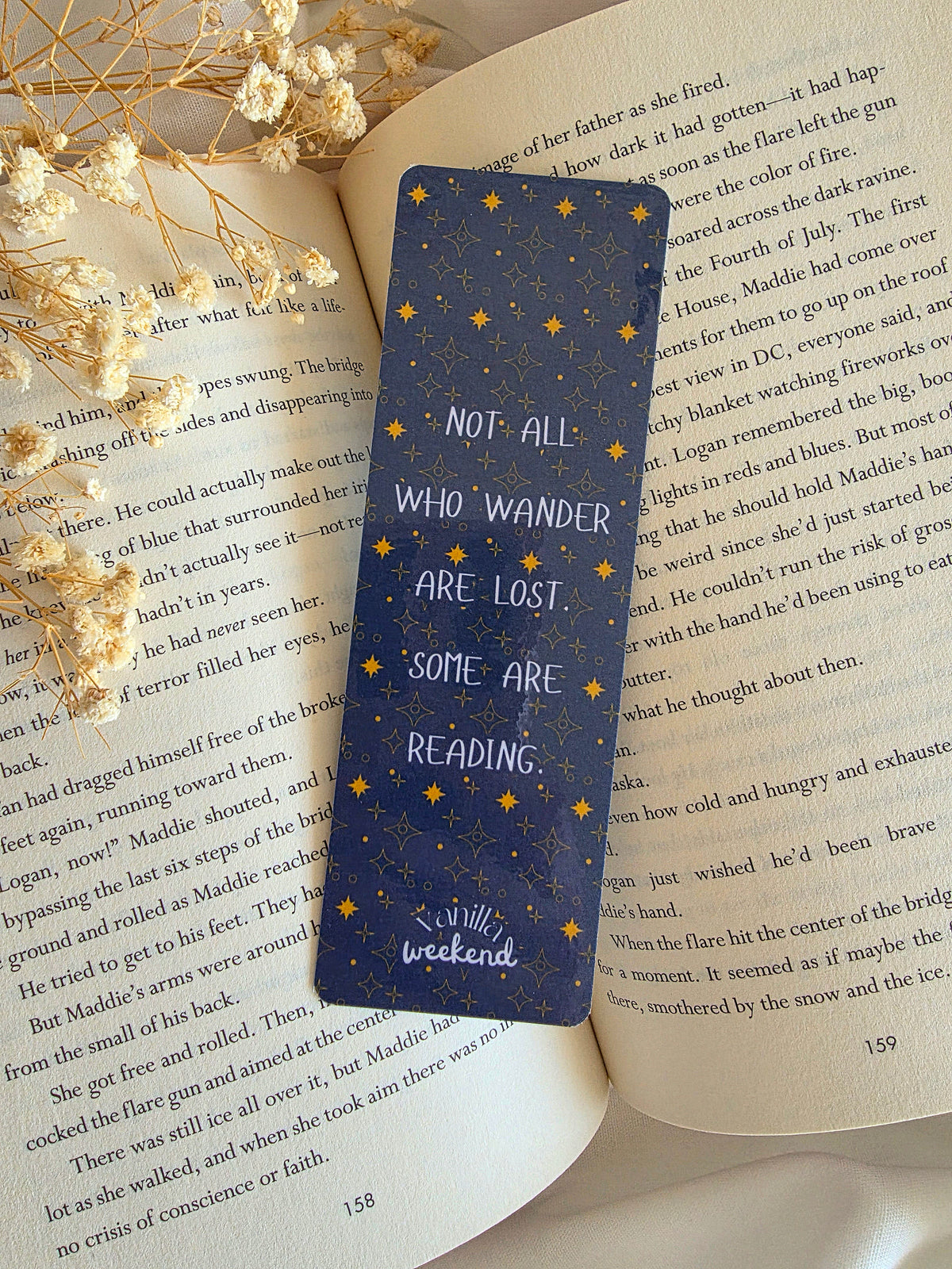 A bookmark with a navy blue background, gold star pattern, and with a text overlay saying "Not all who wander are lost. Some are reading." The bookmark is laying on an open book with baby's breath flowers on the left.