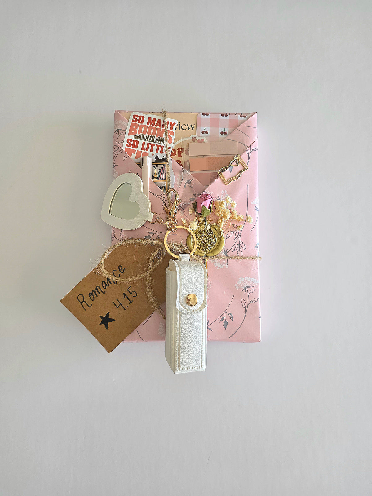 A gently used book wrapped in pink and white floral wrapping paper decorated with a white faux-leather chapstick / lipstick keychain, heart-shaped mirror, and a brown paper tag reading 'Romance 4.15', set against a plain light gray background. Decorated with white and pink flower embellishments wax-sealed to the wrapping paper. Includes a book shaped gold paperclip, stickers, retractable pen, sticky notes, bookmark, and book review card.