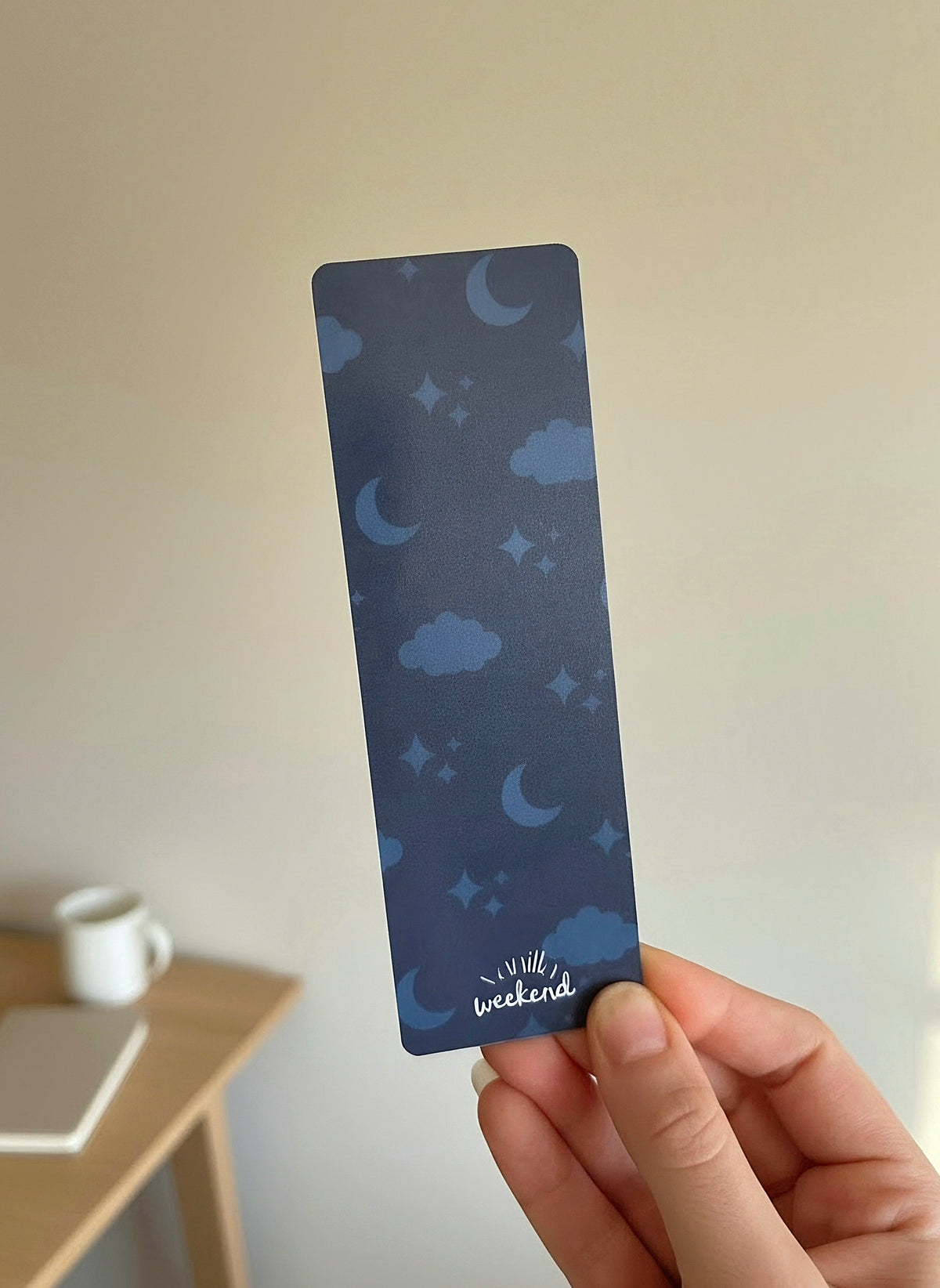 A hand holding a dark navy blue bookmark with a pattern of the moon, stars, and clouds in the background. The words Vanilla Weekend are in the bottom in white.